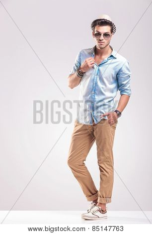 Full length picture of a young casual man posing on grey studio backgroud, holding one hand in his pocket while fixing his collar.