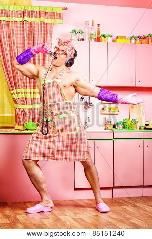 Funny and handsome muscular man in an apron and headphones singing into a microphone in the pink kitchen. Love concept. Valentine's day. Women's day.