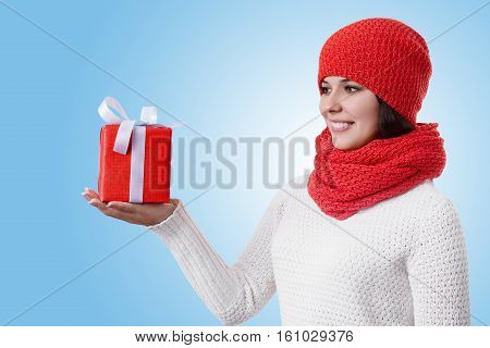 A portrait of an attractive young woman standing sideways on blue background wearing warm winter clothes holding a present in her hand being happy from coming holidays. Receiving New Year presents.