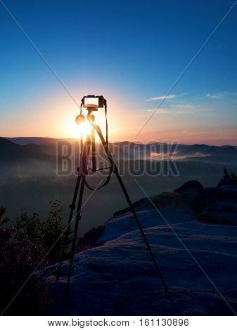 Tripod with running camera on peak ready for photography. Sharp autumn rocky peaks increased from gold fog. Vivid and strong vignetting effect.