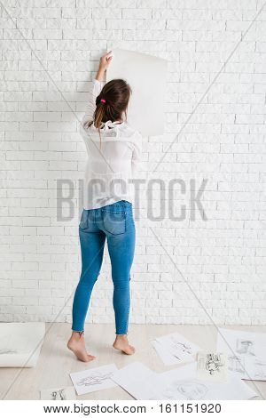 Back of woman hanging empty paper on wall mockup. Artist holding white whatman against white brick side of workshop, free space for text or advertisement