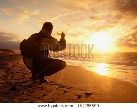 Man Takes Photo Of  Evening Sea. Hiker With Backpack With Phone In Hand. Tourist Squatting