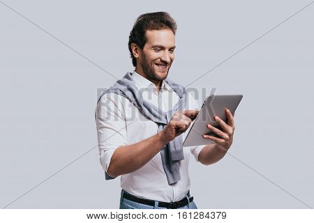 Using modern technologies. Beautiful young man in smart casual clothes working on digital tablet while standing against grey background