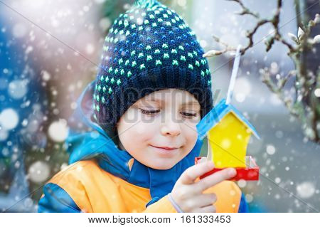 Little kid boy feeding birds in winter. Child hanging colorful selfmade bird house on tree on frosty cold day. Happy preschooler in colorful wam clothes. Selective focus on child and feeder with seeds
