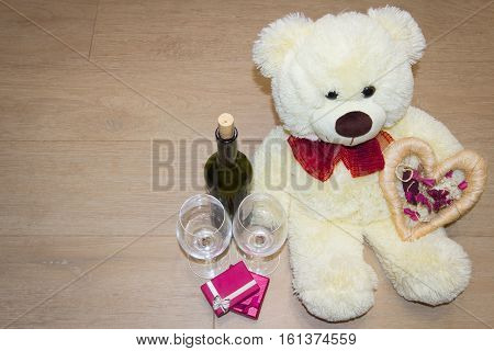 Teddy bear with gifts and love heart, and near glasses and a bottle of wine.