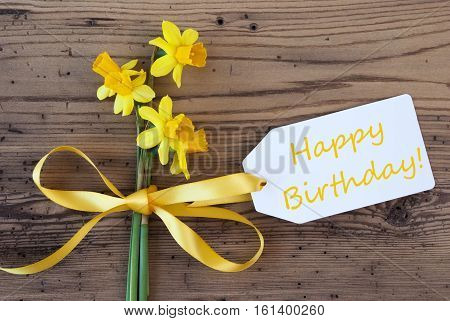 Label With English Text Happy Birthday. Yellow Spring Narcissus Or Daffodil With Ribbon. Aged, Rustic Wodden Background. Greeting Card For Spring Season