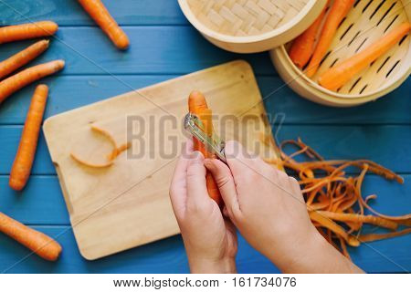 female hands peeling an orange carrot on blue background. Concept of healthy food preparation. Peeled carrots in steamer. Hands in frame. Top down view. Flat lay.