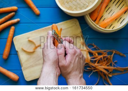 Male hands peeling an orange carrot on blue background. Concept of healthy food preparation. Peeled carrots in steamer. Hands in frame. Top down view. Flat lay.