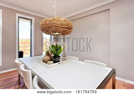 Dining table and chairs are white color and classic the walls are gray in color and there are white color frames around the windows. There is an interior decoration lamp made of bamboo or rattan hanging over the table 