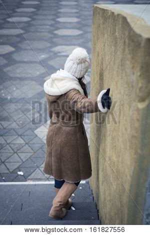A young woman wearing a winter sheepskin coat is standing on some stairs outside by a rock wall.