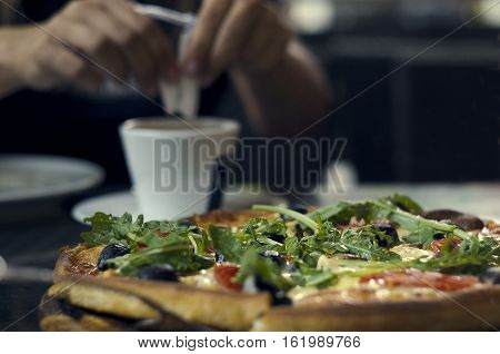 Pizza is on a table in the cafeteria. The girl pours sugar into a cup of coffee in the background.