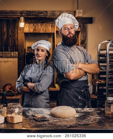 Chef With His Assistant In Cook Uniform Posing With Crossed Arms Near Table With Ready Dough In The 