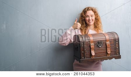 Young redhead woman holding vintage chest happy with big smile doing ok sign, thumb up with fingers, excellent sign