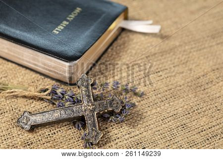 Closeup Of Metalic Christian Cross On Lavanda Flowers Next To Holy Bible On Textile Background.
