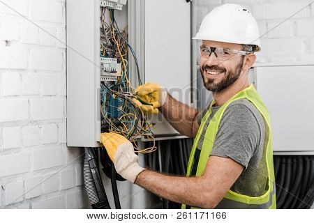 Smiling Handsome Electrician Repairing Electrical Box With Pliers In Corridor And Looking At Camera
