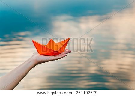 A Young Girl Holds A Paper Boat In Her Hand Above The River. Origami In The Form Of A Ship Has An Or