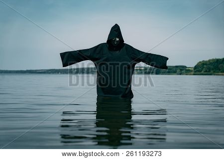 A Man In A Black Robe Stands In The Lake With Hands Outstretched And Looks At The Water On The Backg