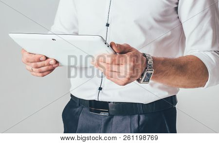 A White Tablet Is In The Hands Of A Man. He Stands In A White Shirt And Black Trousers And Reads Inf