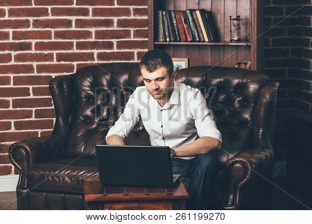 A Man Sits On A Luxurious Sofa And Works Behind A Laptop In His Office On The Background Of The Book