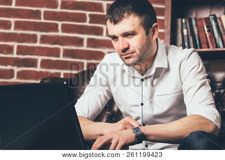 Handsome Man Looks At The  Laptop Screen On Background Of Office Cabinet. He Is Dressed In The Busin