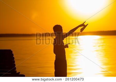 Little Boy Stands On The Background Of The River At Sunset. He Holds A Paper Airplane In His Hand An
