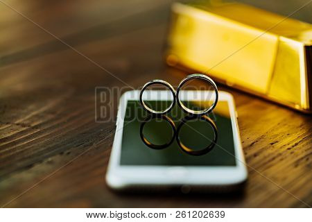 Two Wedding Rings Are On The Phone's Display. A Pair Of Gold Rings Are On The Background Of A Wooden