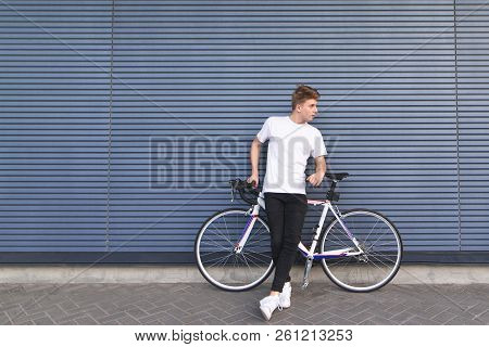 Portrait Of A Young Man In Full Height Standing Beside A White Bicycle Against The Wall And Looking 