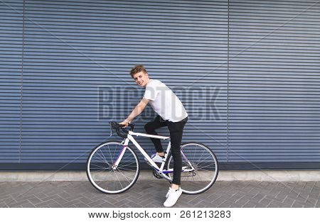 Happy Young Man Sitting On A White Bike Against The Wall, Looking At The Camera And Smiling. Guy Rid