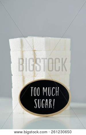 a pile of white sugar cubes, disposed in a block, and a black signboard with the text too much sugar written in it, against an off-white background