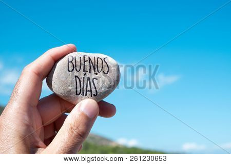 closeup of a young caucasian man showing a stone with the text buenos dias, good morning in spanish written in it, holding it against the blue sky