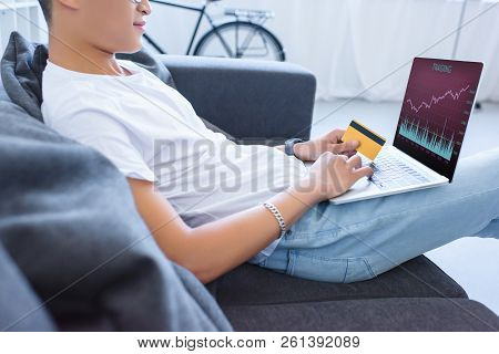 Cropped Image Of Man Using Laptop With Traiding Appliance And Holding Credit Card On Sofa At Home