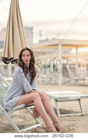 Romantic young beautiful girl with long fair hair in short dress sitting in chaise-longue on the beach. Attractive sleepy woman dreaming at dawn on shore in Italy. Outdoor female portrait, soft focus