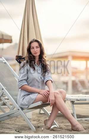 Romantic young beautiful girl with long fair hair in short dress sitting in chaise-longue on the beach. Attractive sleepy calm woman dreaming at dawn on the shore in Italy, Europe. Soft focus
