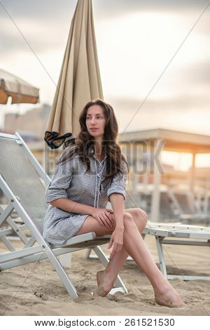 Romantic young beautiful girl with long fair hair in short dress sitting in chaise-longue on the beach. Attractive sleepy calm woman dreaming at dawn on the shore in Italy, Europe. Soft focus