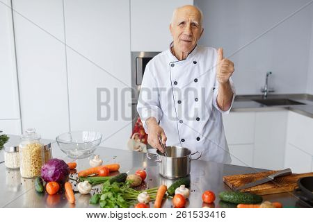 The Aged Chef Man Cooking A Soup And Showing Thumb Up In The Kitchen. The Concepy Of Cooking