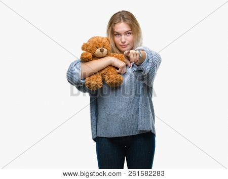 Young caucasian woman holding cute teddy bear over isolated background pointing with finger to the camera and to you, hand sign, positive and confident gesture from the front