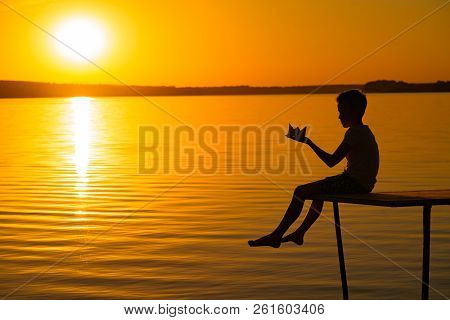 A Little Child Is On A Bridge With Origami In The Shape Of A Boat In His Hands At Sunset. The Legs O
