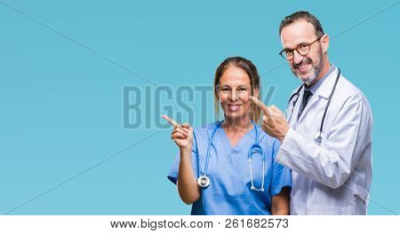 Middle age hispanic doctors partners couple wearing medical uniform over isolated background smiling and looking at the camera pointing with two hands and fingers to the side.