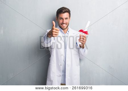 Handsome young doctor man over grey grunge wall holding diploma happy with big smile doing ok sign, thumb up with fingers, excellent sign