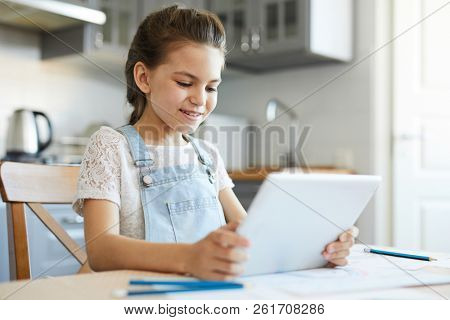 Pretty youngster with touchpad watching vids in the net while sitting in the kitchen