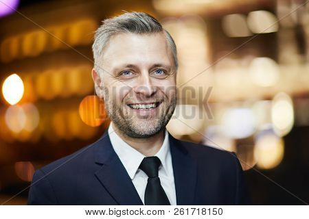 Cheerful man in elegant suit and tie enjoying walk in urban environment aith night lights
