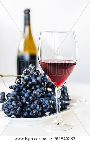 Glass Of Red Wine With A Bottle And A Cluster Of Black Grapes On White Table. Selecive Focus.