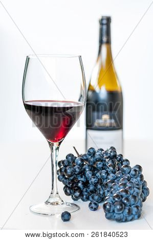 Glass Of Red Wine With A Bottle And A Cluster Of Black Grapes On White Table. Selecive Focus.