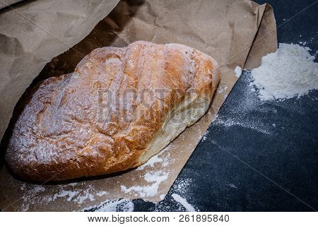 A Loaf Of White Bread On Brown Craft Paper, Powdered With Flour On A Dark Table Background.
