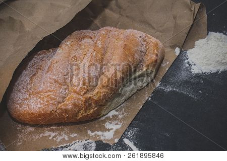 A Loaf Of White Bread On Brown Craft Paper, Powdered With Flour On A Dark Table Background.