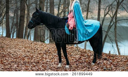 A Girl With Long Hair, Blonde Hair, Sits On The Back Of A Black Horse, In A Blue Cape And A Vintage 