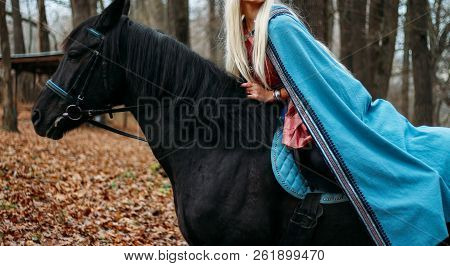 A Girl With Long Hair, Blonde Hair, Sits On The Back Of A Black Horse, In A Blue Cape And A Vintage 