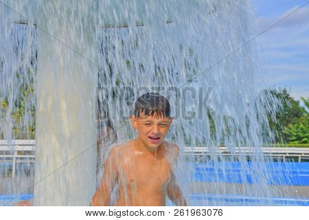 Happy Little Boy Enjoying Summer Day In The Swimming Pool. Boy Standing Under Sprinkler In Spray Poo