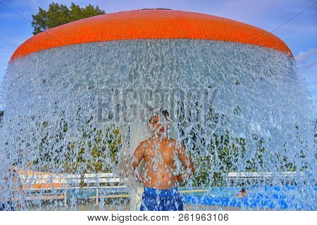 Happy Little Boy Enjoying Summer Day In The Swimming Pool. Boy Standing Under Sprinkler In Spray Poo