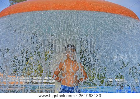 Little Boy In The Small Swimming Pool. Boy Taking Shower In Water Park. Boy Standing Under Sprinkler